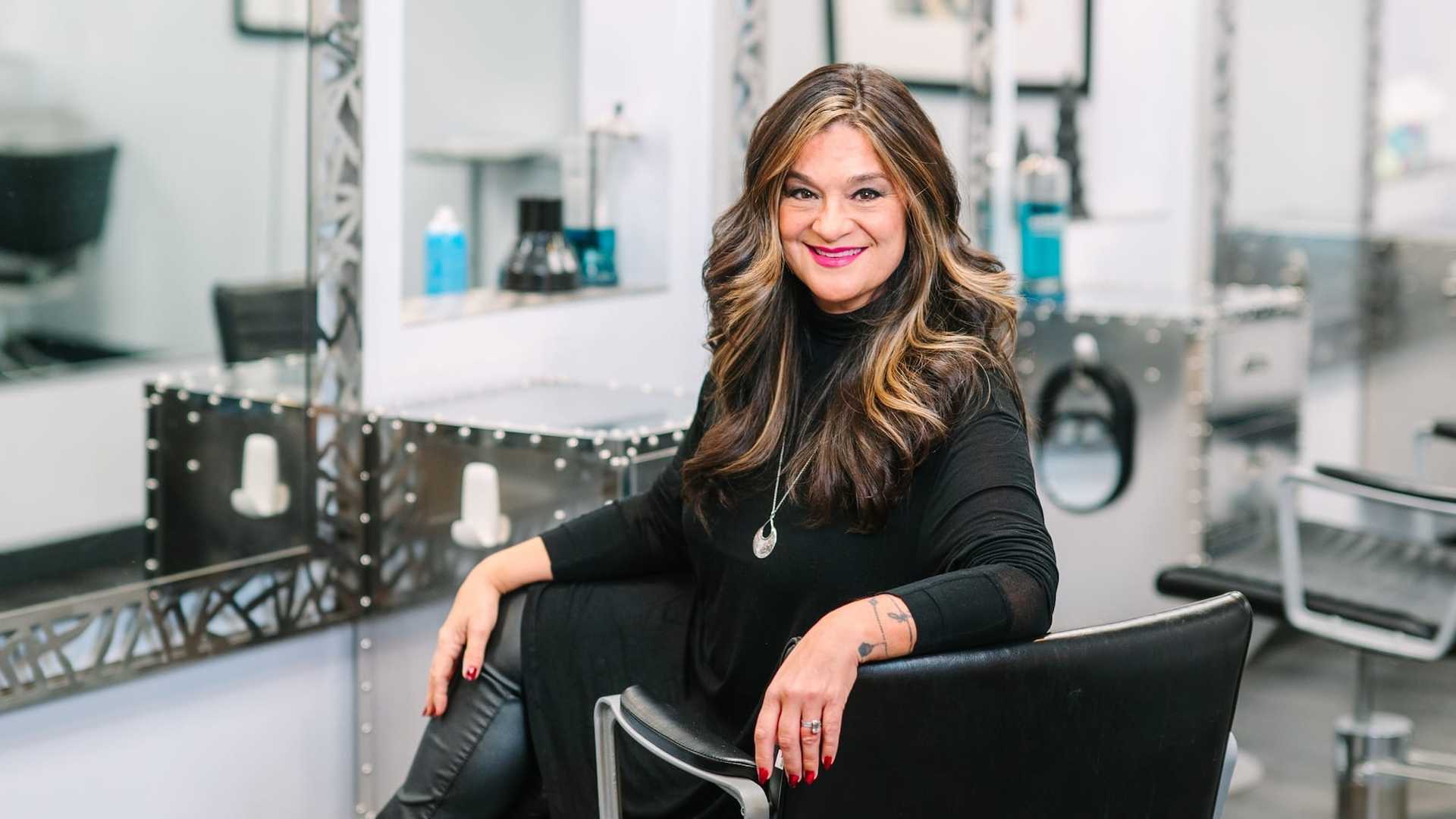 Woman in a salon, smiling while seated, with salon equipment in the background.