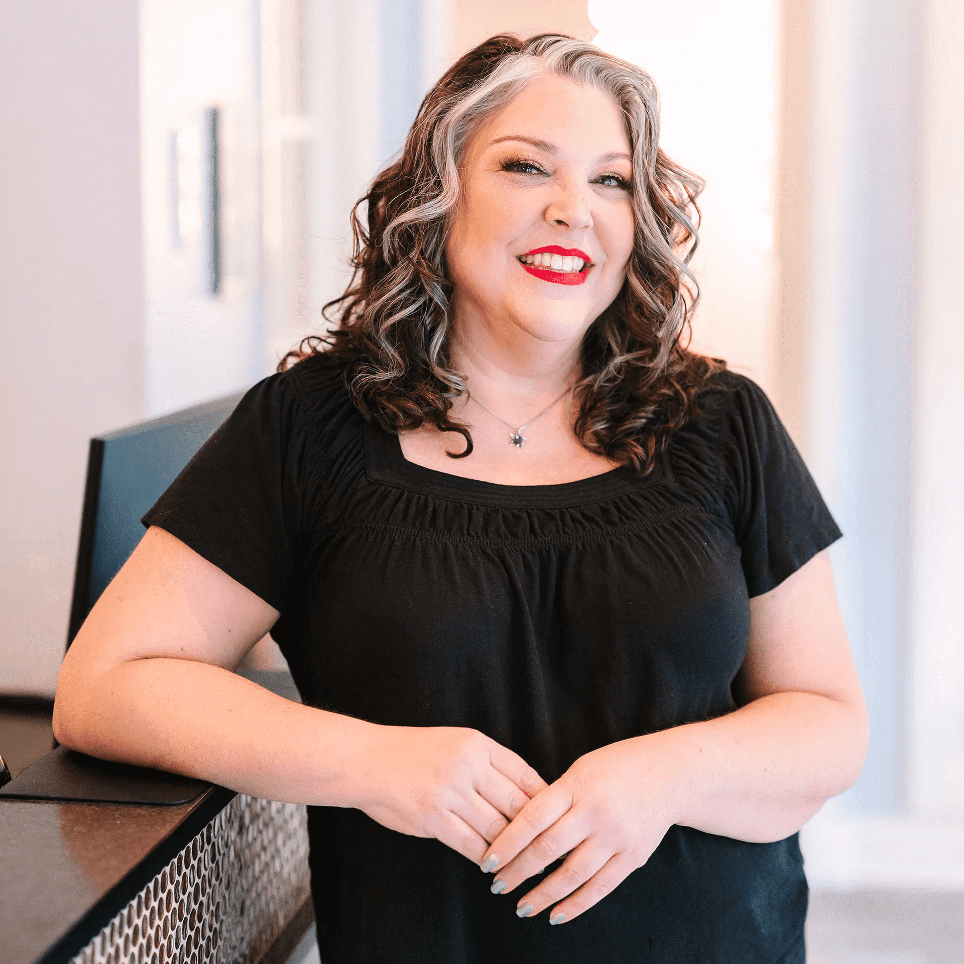 Smiling woman with wavy hair and red lipstick, wearing a black top, leaning on a counter.