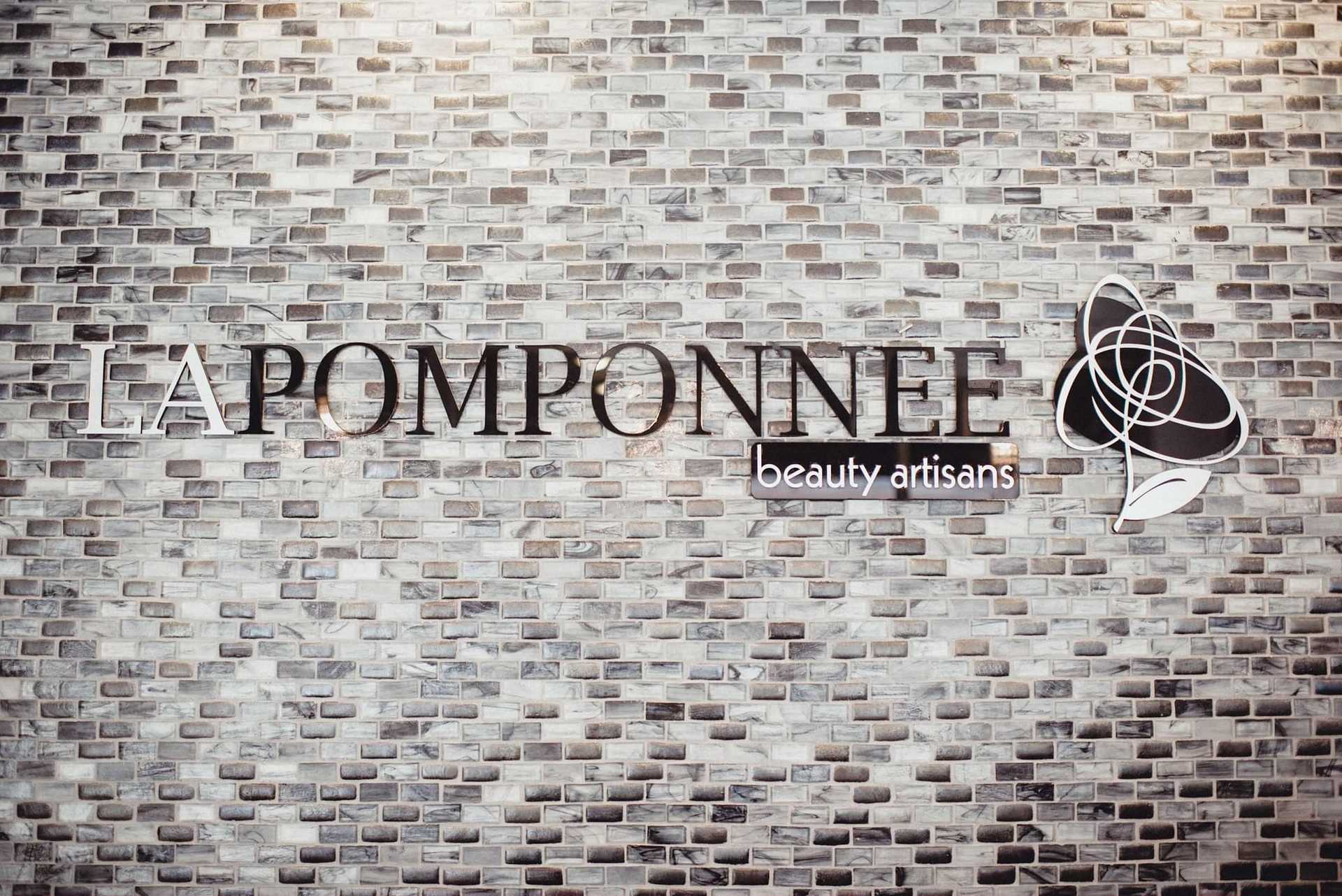 Sign for "LA POMPONNEE beauty artisans" on a brick wall background.